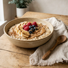 A beige speckled ceramic bowl filled with oatmeal topped with banana slices, peanut butter, raspberries, and blueberries, set on a rustic wooden table next to a linen napkin and wooden spoon.
